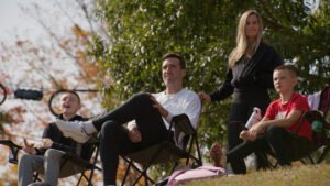 "A family of four, two adults and two kids, sit outdoors on folding chairs on a hillside, enjoying a sunny day with trees and autumn foliage in the background."