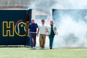 "Three men walk confidently out of stadium tunnel onto grass field, one wearing sunglasses, smoke or fog surrounds them, colorful sign with chicken graphic and large yellow letters in background, midday outdoor setting."