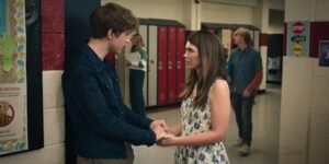 "A teenage boy and girl hold hands in a high school hallway near lockers, facing each other and talking seriously. Other students and colorful posters are visible in the background, creating a typical school setting."