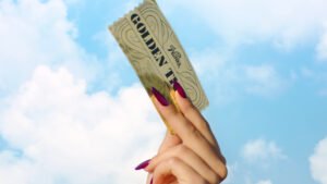 "A hand with long, painted nails holds up a golden ticket against a blue sky with clouds."